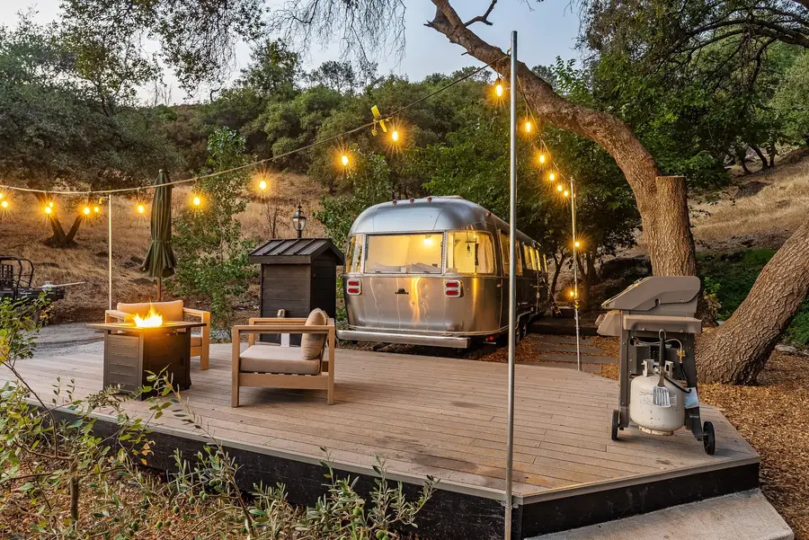 The wooden deck of Airstream 1A at dusk, with a propane fire pit, chairs and string lights under the oaks
