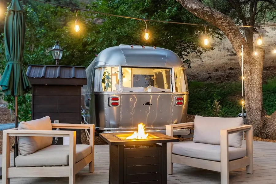 An Airstream's outdoor living space with lounge chairs, a lit propane fire pit and string lights