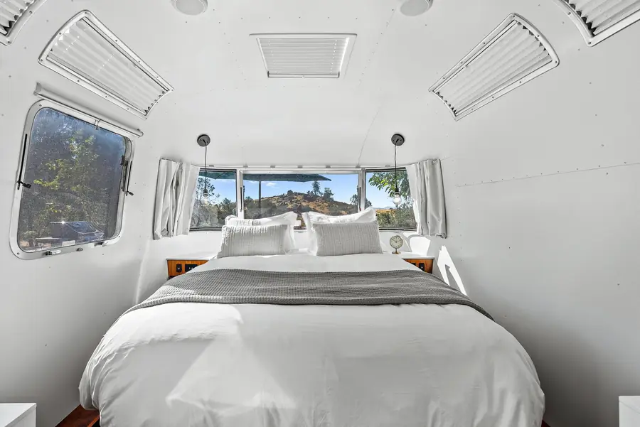 Queen bed in white linens at the back of the Airstream, with windows on three sides looking out to the hills