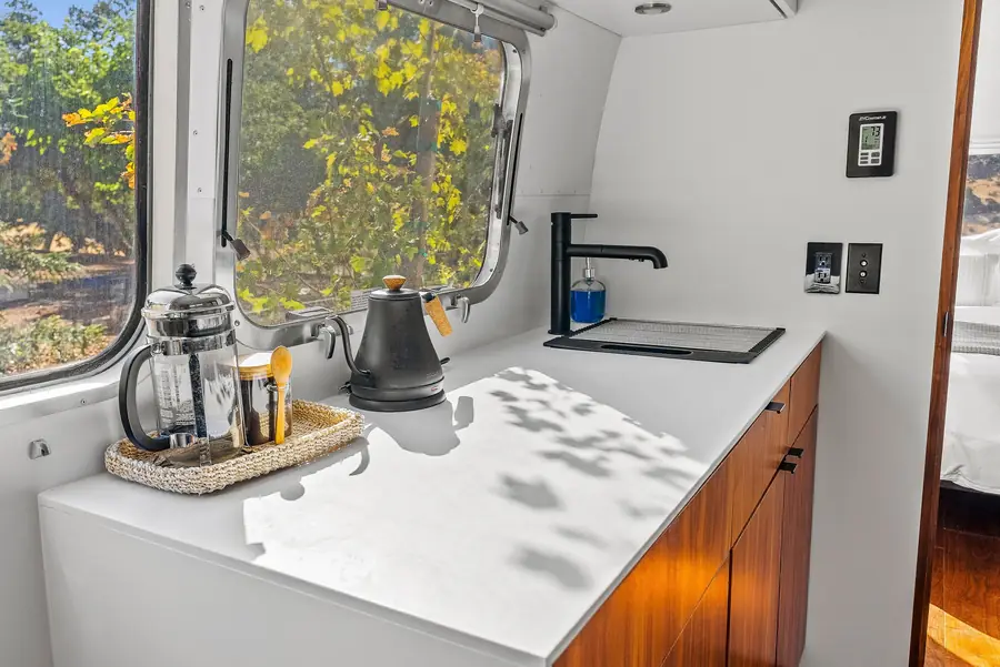 Airstream kitchenette with a French press, kettle and coffee tray on a white counter below a window to the oaks