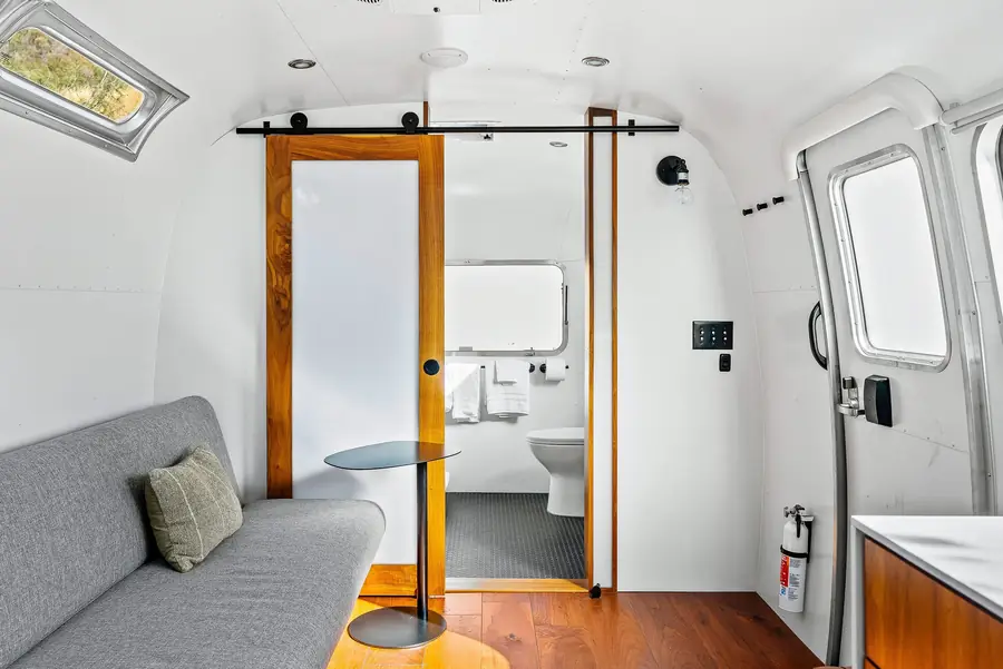 Airstream living area with a gray pull-out futon and a small round table, facing a walnut barn door to the bathroom
