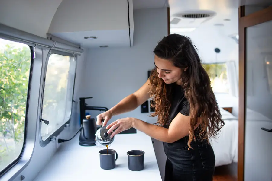 A guest pours coffee at the kitchenette counter, with windows to the oaks behind her