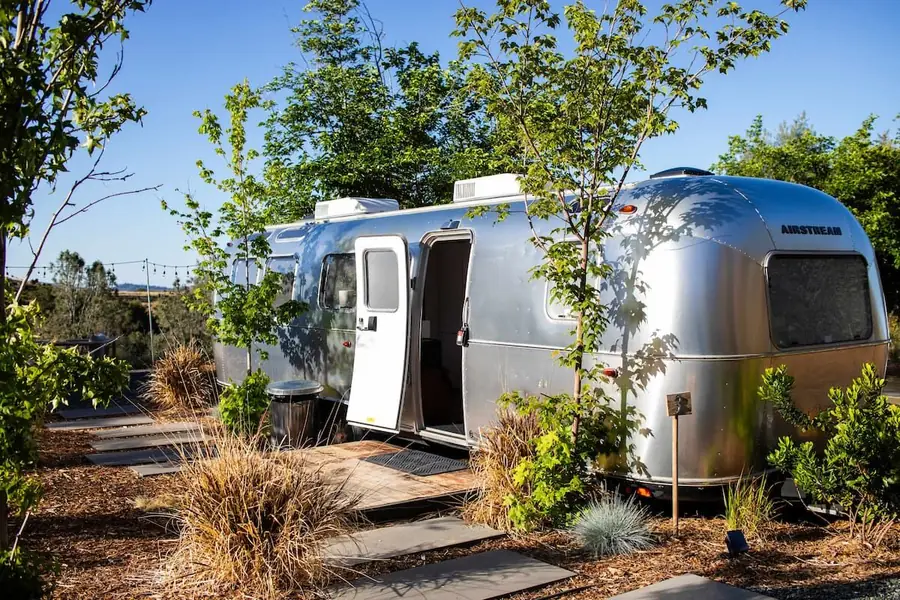 Airstream with its door open, among young oaks and a stone path