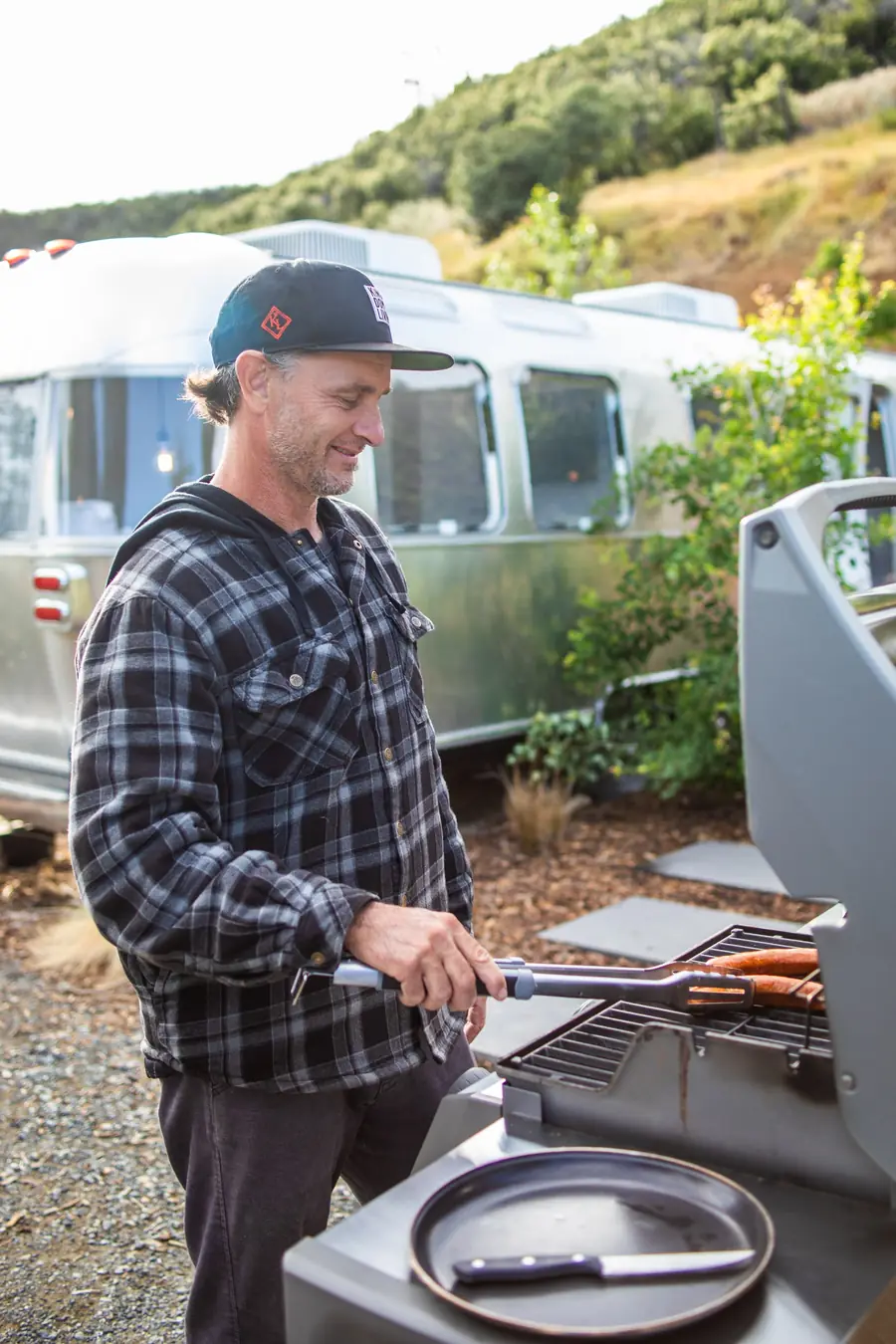 A man grilling sausages on a BBQ grill beside an Airstream