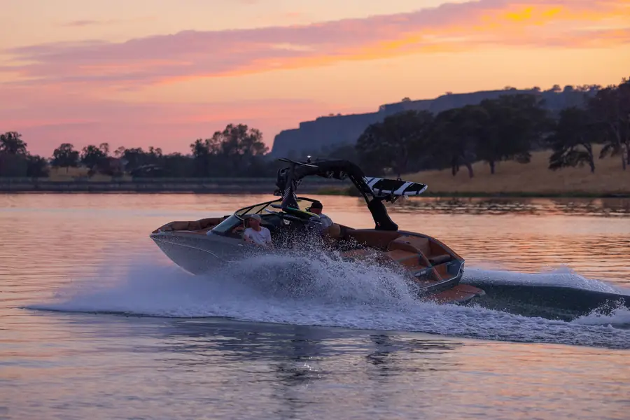 A wake boat cruising across a calm lake at sunset under a pink sky