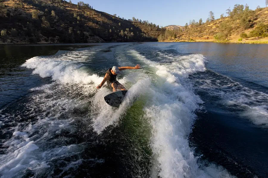 A wakeboarder cutting across a white boat wake on a foothill lake