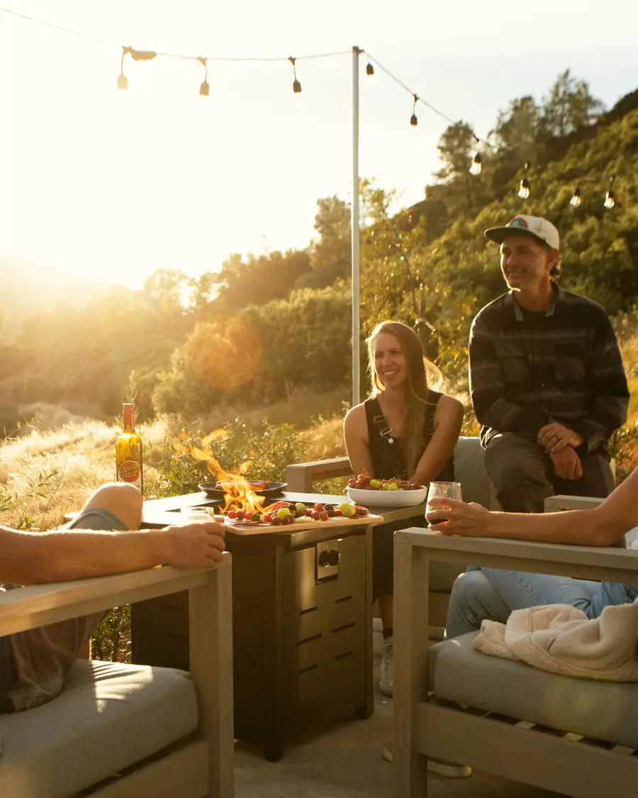 Friends talking around a propane fire table outside their Airstream at sunset, string lights overhead