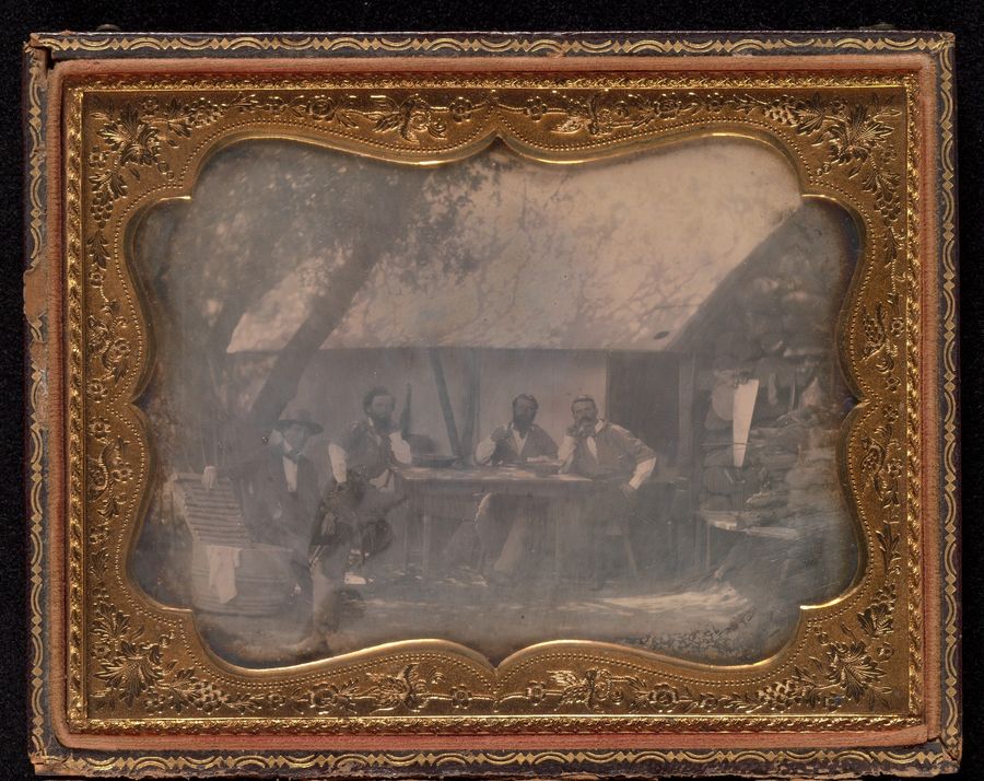 Daguerreotype in a brass mat of four gold miners seated in front of their wooden house
