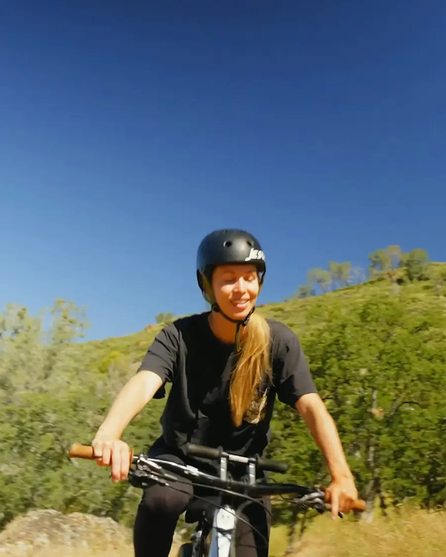 A smiling woman in a helmet riding a mountain bike toward the camera, green hills behind