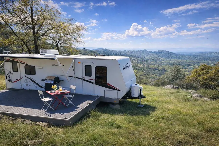 The white Mariposa RV on its own deck with a small table and chairs, looking out over oak-covered foothills
