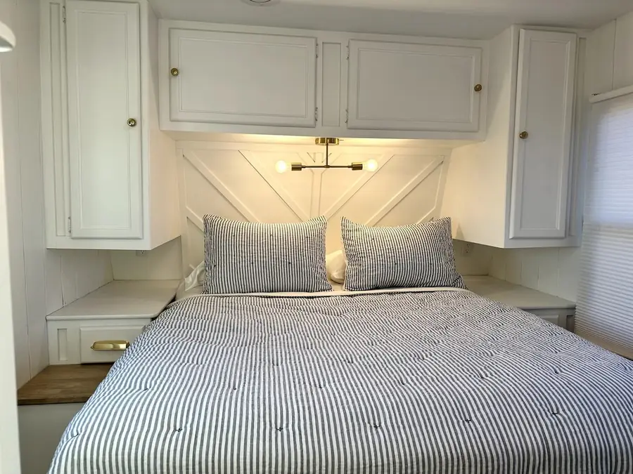 The bed in the Mariposa RV with striped bedding, white cabinets and a wall light